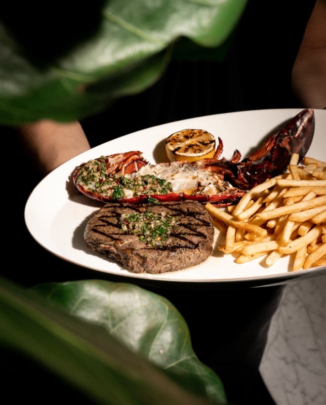 A white plate with steak, lobster, grilled lemon, and French fries