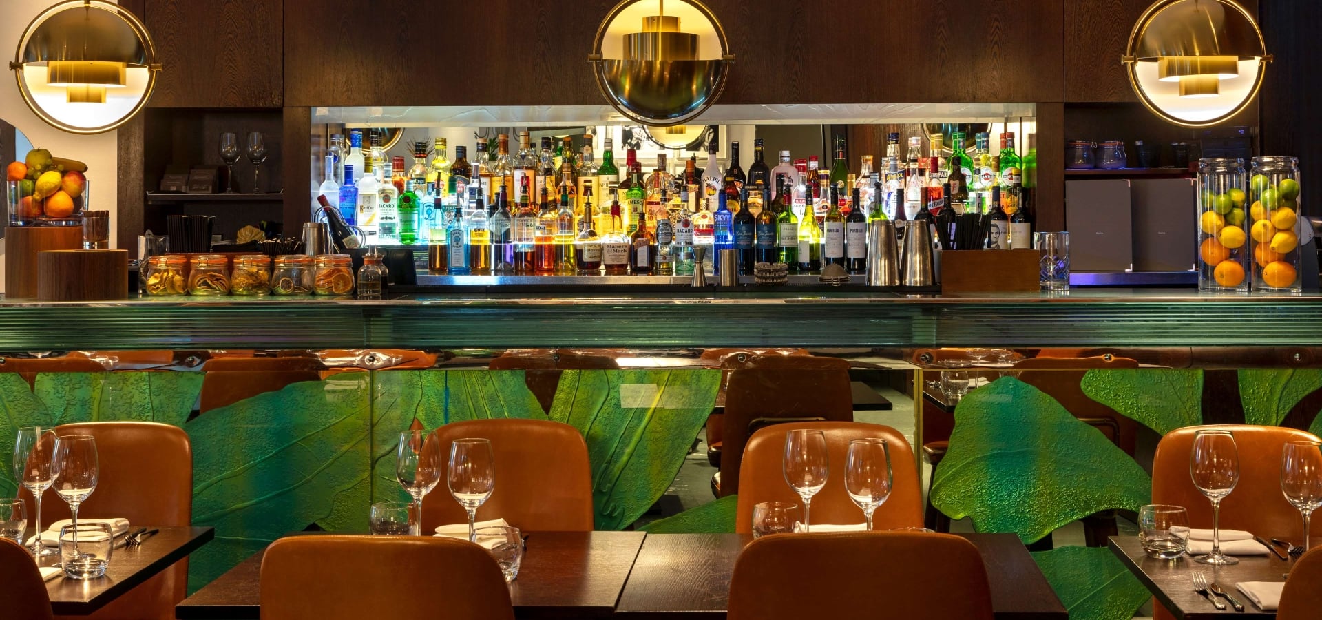 Bar with backlit spirits display, tan leather brown chairs, tropical leaf panelling, and gold pendant lights.