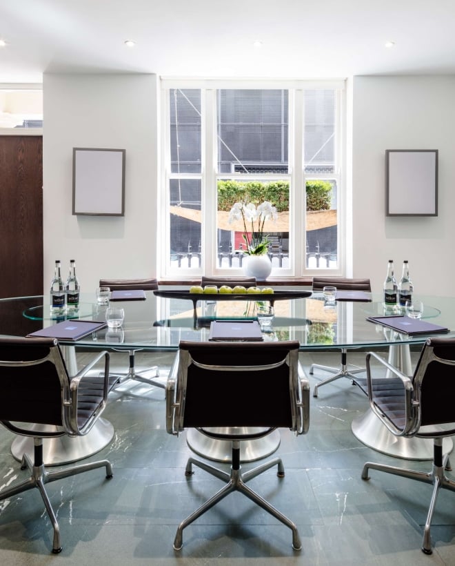 Private meeting room with oval glass table, leather office chairs, wall-mounted TV, and natural light from floor to ceiling windows.