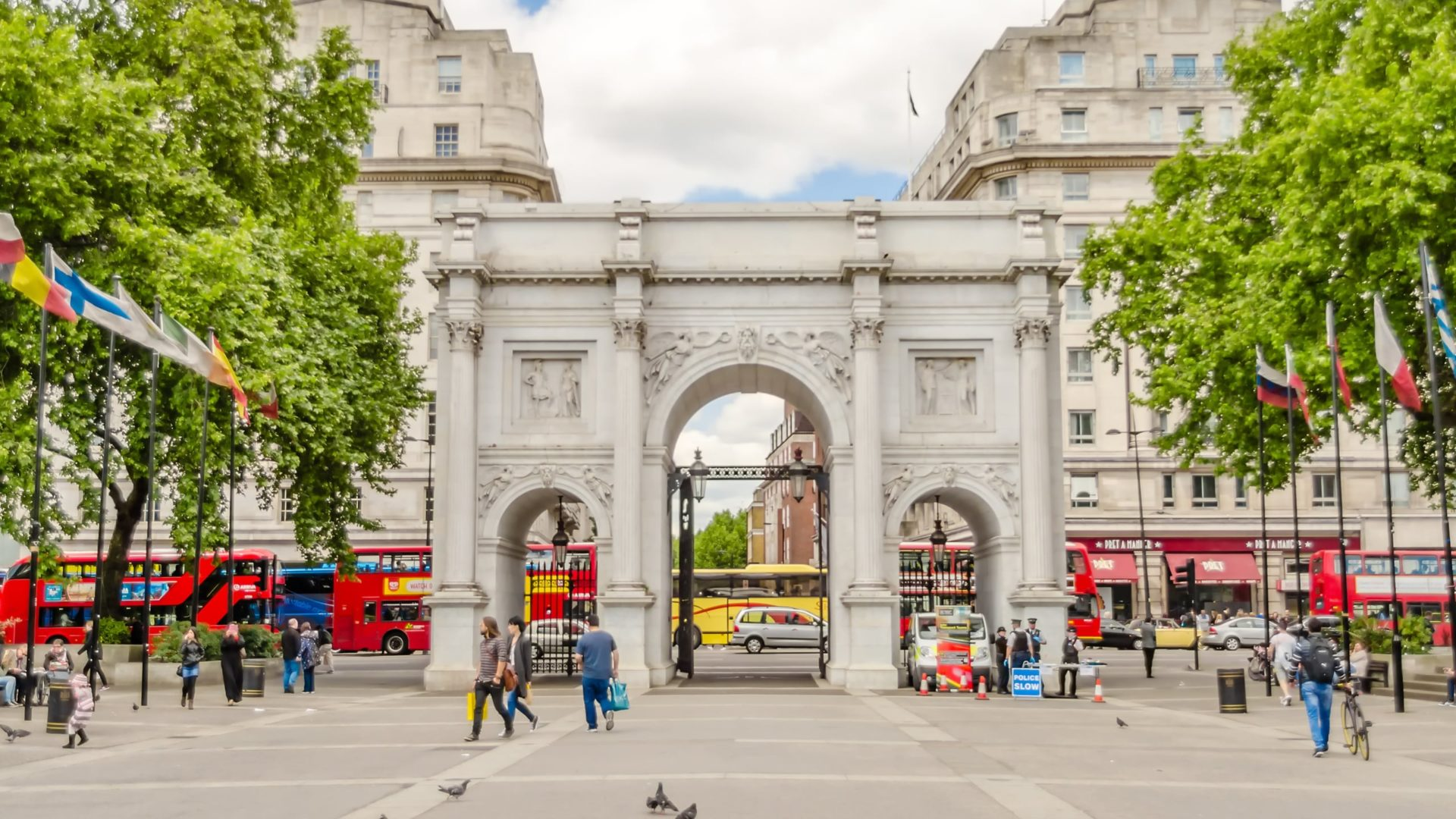 Marble Arch landmark in London with red double-decker buses, international flags, pedestrians, and green trees.