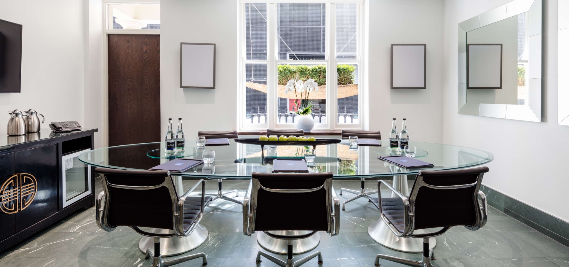 Private meeting room with oval glass table, leather office chairs, wall-mounted TV, and natural light from floor to ceiling windows.